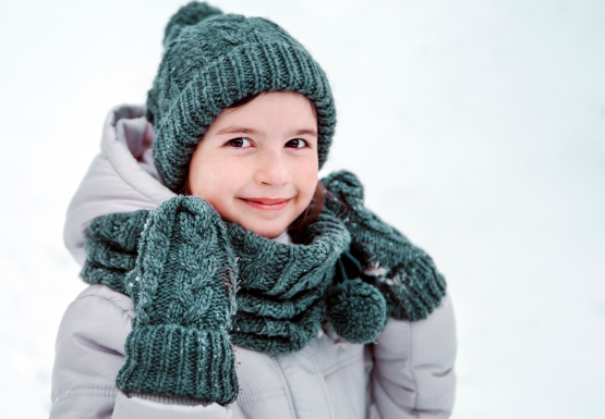 Kind ist eingepackt in Winterbekleidung. Es trägt eine graue Winterjacke eine tannengrüne Mütze, Handschuhe und Schal.