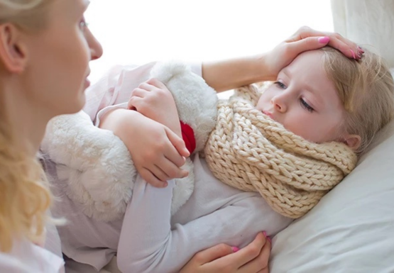 Kind hat Halsschmerzen und liegt mit dickem Schal und Teddybär im Arm im Bett. Die Mutter fasst ihrer Tochter liebevoll an den Kopf.